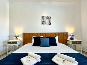 Ein ordentlich bezogenes Bett mit einem Holz-Kopfteil, zwei Nachttischen mit Lampen und einem Kunstwerk darüber.