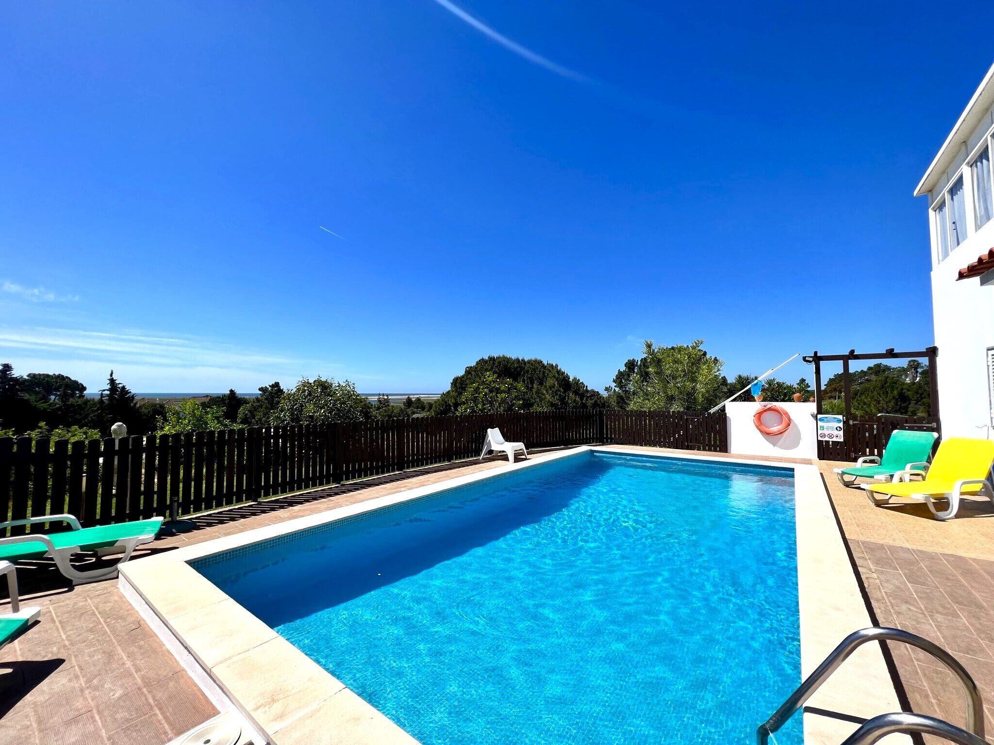 Ein rechteckiger Swimmingpool mit glasklarer blauer Wasseroberfläche, umgeben von einer gefliesten Liegefläche. Mehrere Liegestühle und eine Poolleiter sind sichtbar.