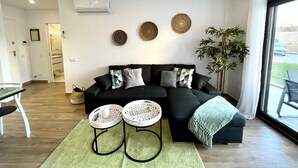Un salon moderne avec un canapé d’angle noir, deux petites tables d’appoint blanches, un tapis vert et une grande fenêtre donnant sur l’extérieur.