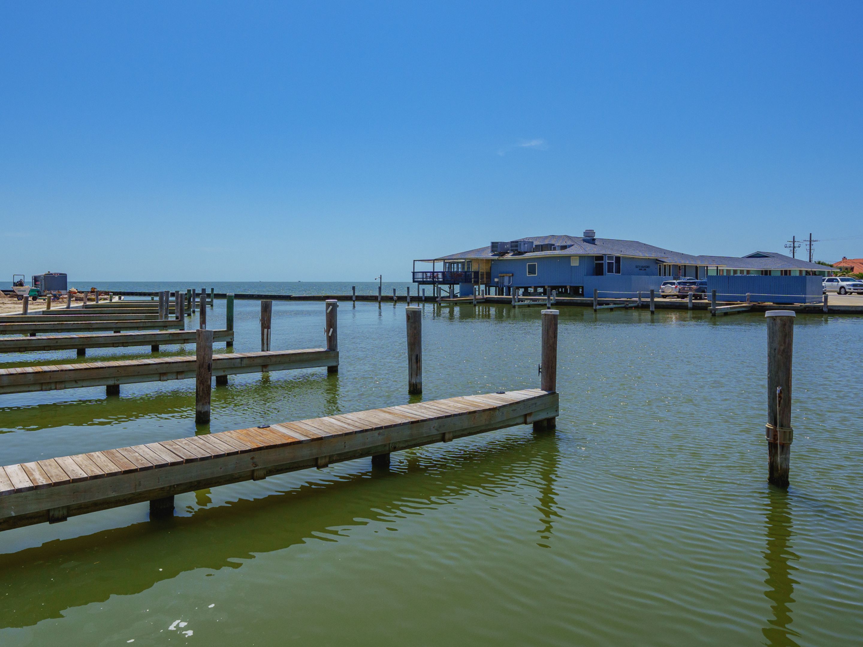A waterfront property with multiple wooden docks extending into the water, a large blue building with a flat roof, and a clear blue sky.