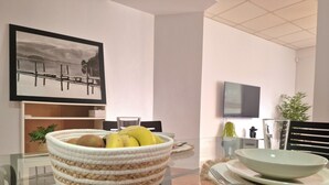 A dining area with a glass table, chairs, a woven basket with fruits, a framed picture on the wall, and a television mounted on the wall.