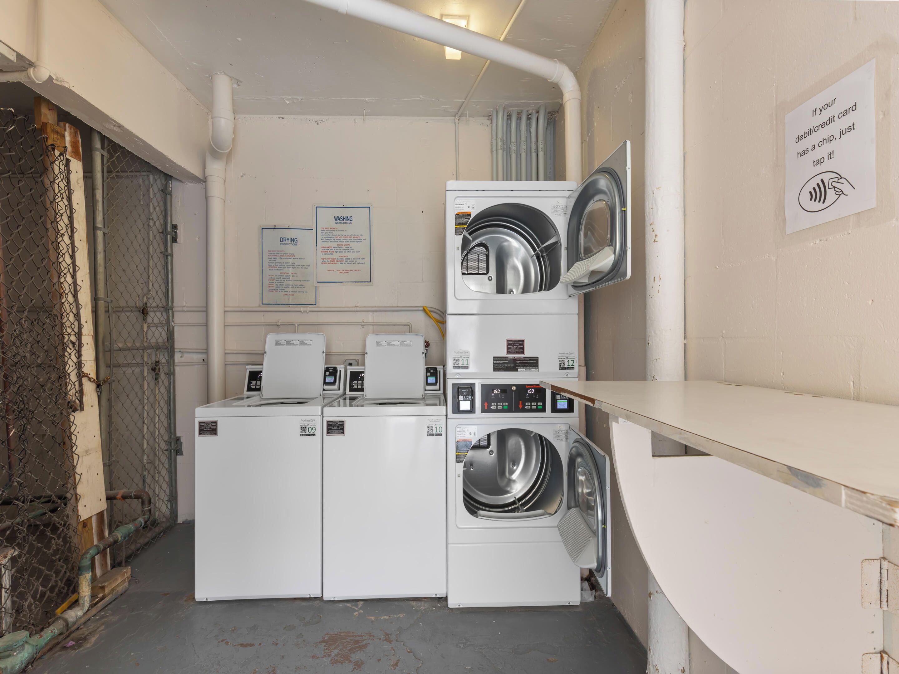Un espace buanderie avec des machines à laver et des sèche-linge superposés, un panneau indiquant les instructions de lavage et de séchage, et un avis concernant le paiement par carte.