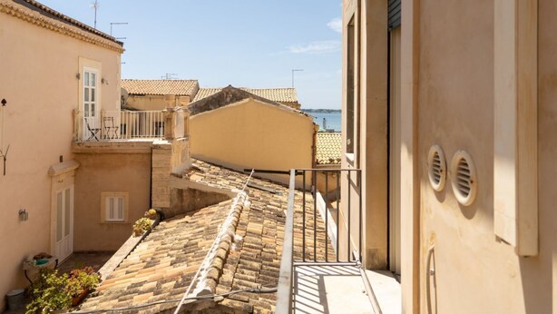 Una calle estrecha con techos de tejas de terracota, un pequeño balcón con barandilla y un edificio de fachada color crema.