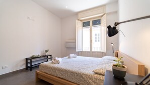 Un dormitorio con cama, mesita de noche, lámpara, planta en maceta y ventana con cortinas.