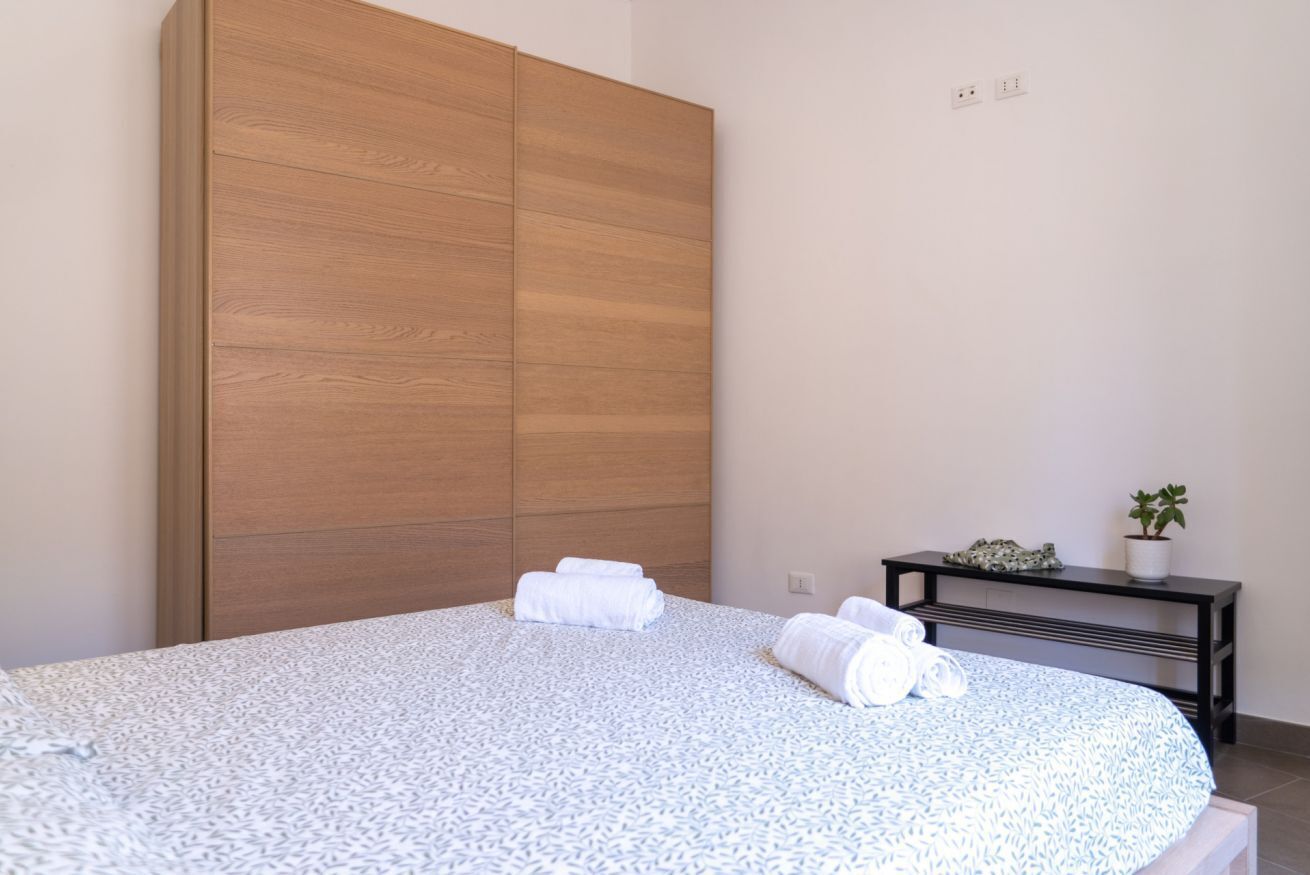 Un dormitorio con una cama, un armario de madera, una mesita con una planta y un enchufe de pared.