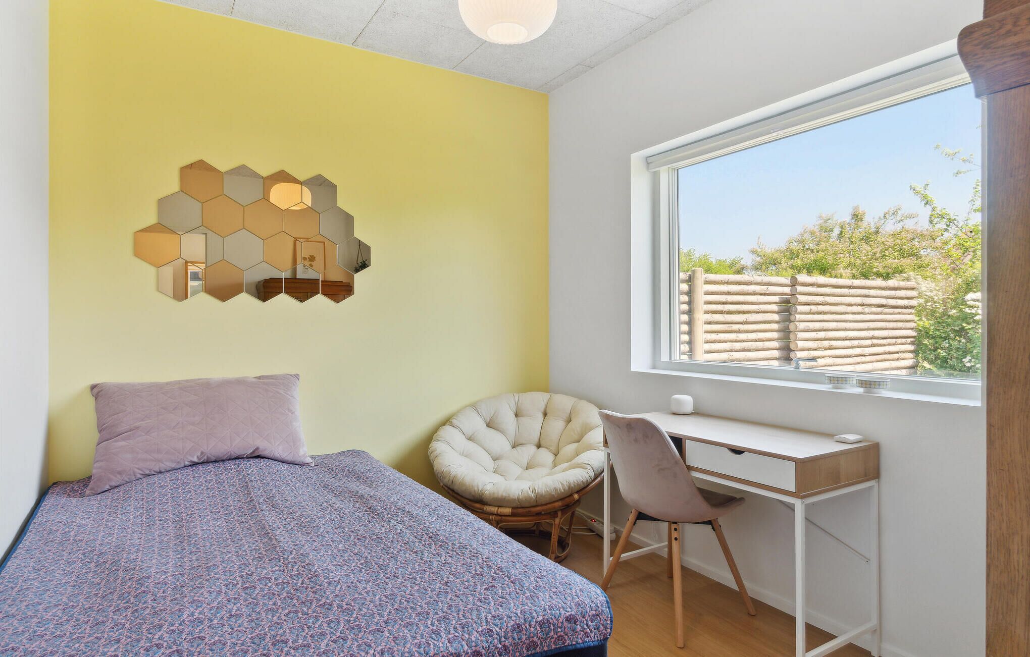 Een slaapkamer met een bed, een bureau, een stoel, een raam en een zeshoekige spiegel aan de muur.