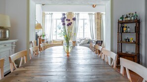 Ett matrum med ett träbord, stolar, en vas med blommor, en lampa och en hylla med flaskor.