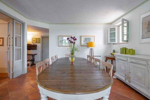 Un comedor con una mesa de madera, sillas, un florero con flores, cuadros enmarcados, una lámpara y un armario.