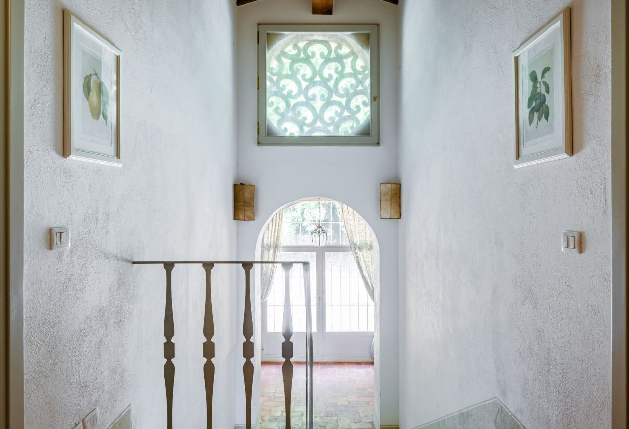 A hallway with a staircase, framed artwork, a decorative window, and a door leading to an outdoor area.