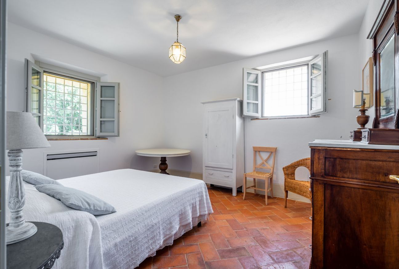 Un dormitorio con una cama, una mesa redonda, una silla, un armario y dos ventanas.