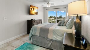 A bedroom with a bed, a TV mounted on the wall, a ceiling fan, a nightstand with a lamp, and a view of the outdoors through a sliding door.