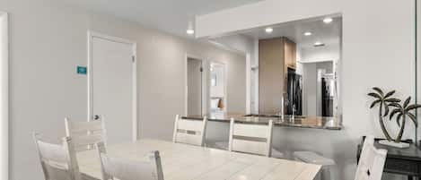 A modern dining area with a rectangular table and six chairs, a kitchen with a refrigerator and stove, and a large mirror above the counter.