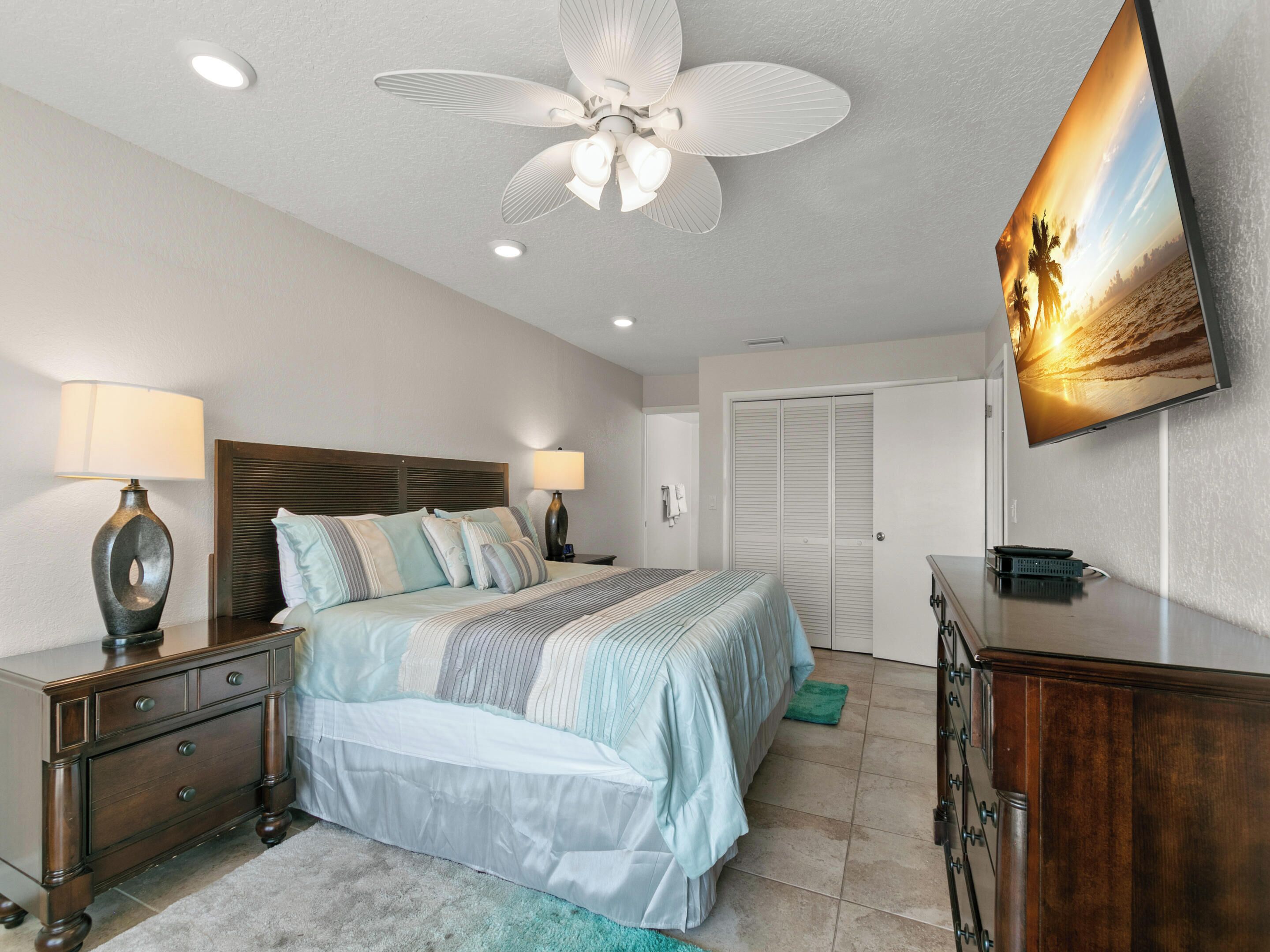 A bedroom with a bed, nightstands, a dresser, a ceiling fan, recessed lighting, a TV, and a door.