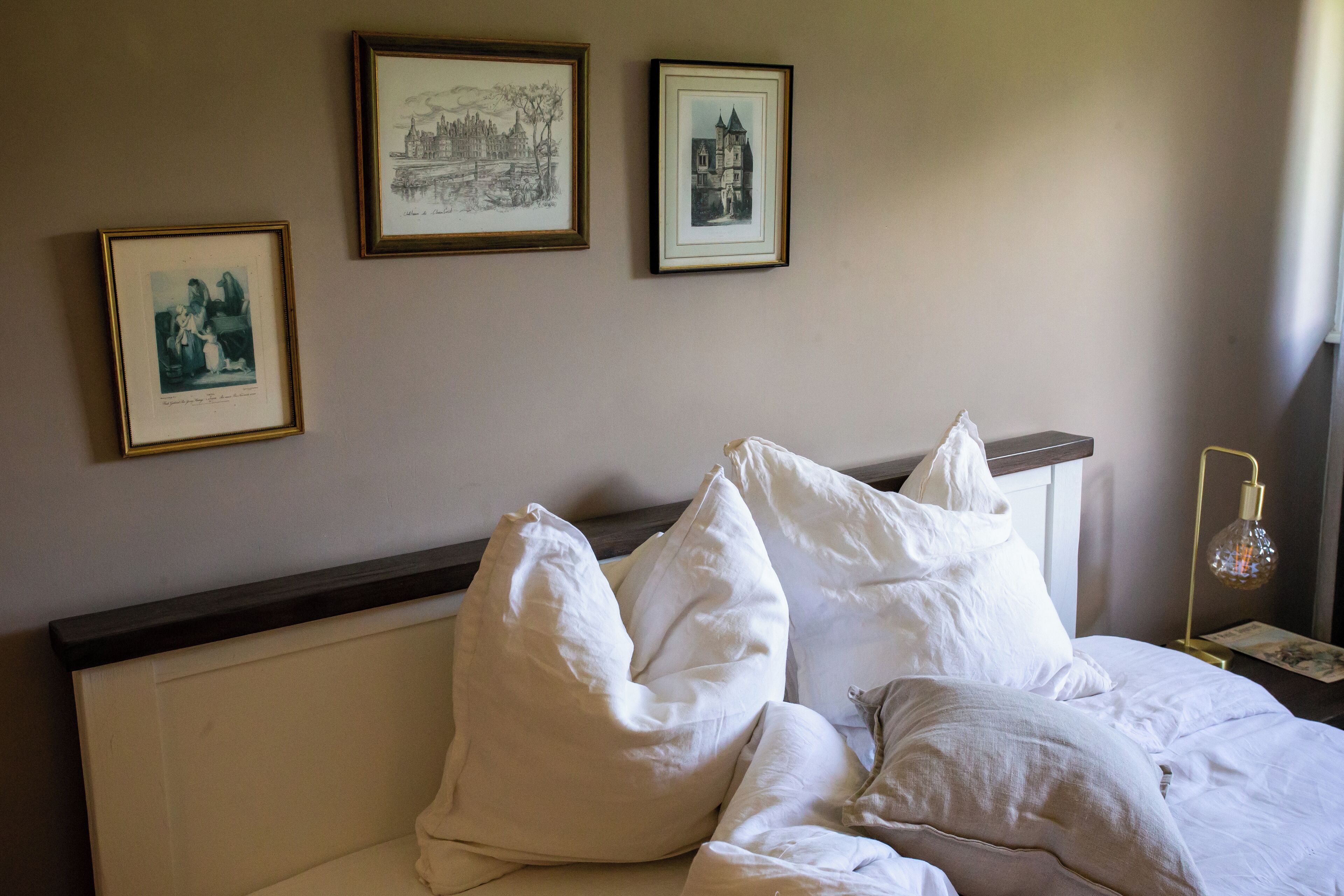A neatly made bed with white bedding and pillows, a framed picture of a castle on the wall, and a lamp on the nightstand.