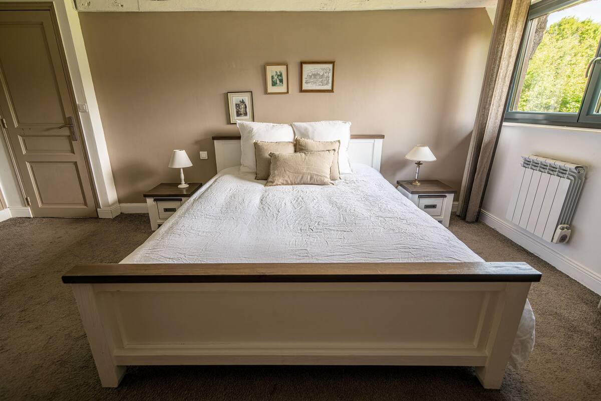 Un dormitorio con una cama grande, dos mesitas de noche con lámparas, un radiador y cuadros enmarcados en la pared.