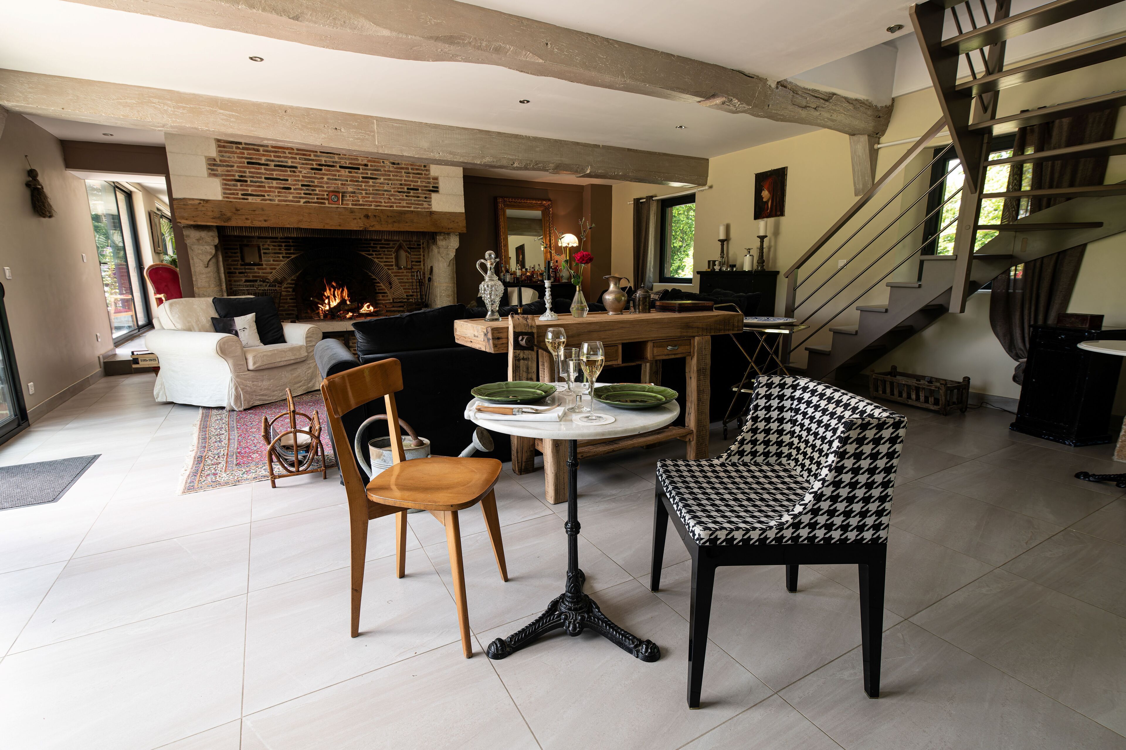 A spacious living area with a fireplace, a dining table with chairs, a houndstooth patterned chair, and a staircase.