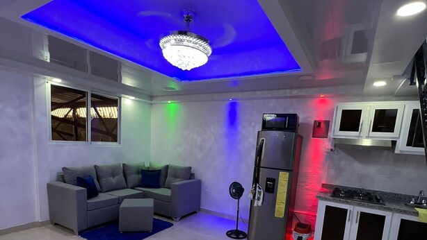 Ein modernes Wohnzimmer mit blau beleuchteter Decke, einem grauen Sofa, einem kleinen Ventilator und einer Küchenzeile mit Schränken und einem Kühlschrank.