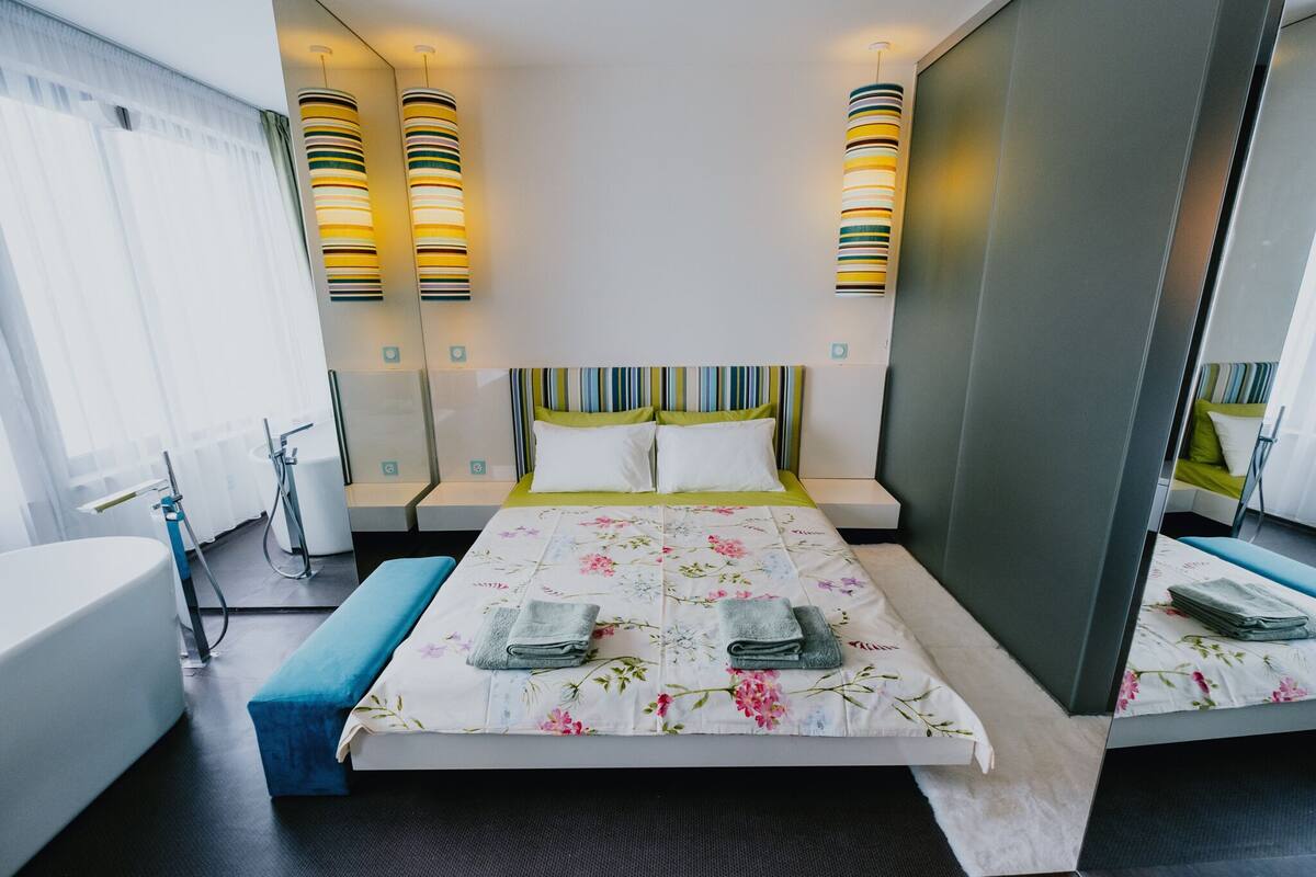 A modern hotel room with a large bed, two bedside tables, a built-in bench, a large window with curtains, and a bathroom with a bathtub and shower.