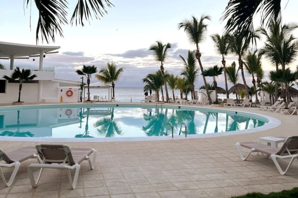A swimming pool with lounge chairs, palm trees, and a view of the ocean.
