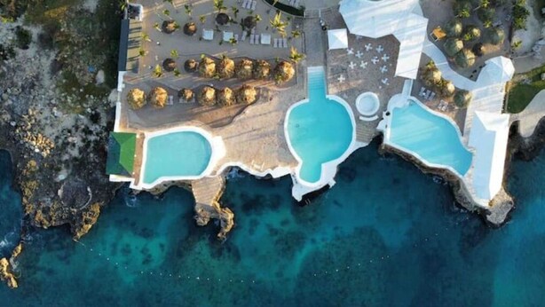 An aerial view of a resort with two large swimming pools, a beach area with lounge chairs, and a rocky shoreline.