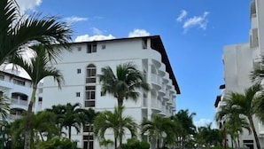 A white multi-story hotel with balconies, surrounded by palm trees and a paved walkway.