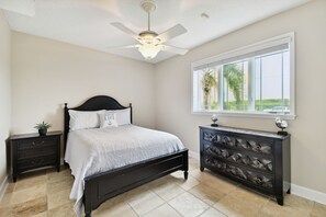 A bedroom with a bed, a nightstand, a dresser, a ceiling fan, and a window with a view of palm trees.