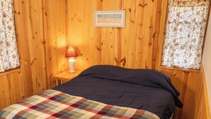A wooden-paneled room with a bed, a nightstand, and a lamp. Text advises guests to bring their own bedding and towels.