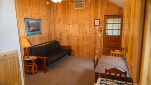 A cozy room with wooden walls, a sofa, a small table with a lamp, a chair, and a window with curtains.