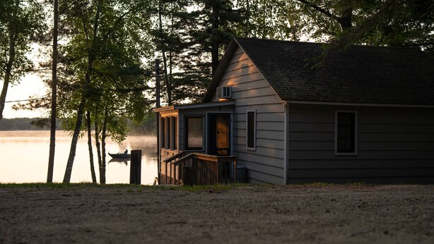 A small cabin with a porch, located near a body of water and surrounded by trees.