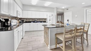 A modern kitchen with white cabinetry, a central island, and a dining area with wooden chairs. - Admirals Quarters 802 - Coastal Luxury with Endless Gulf View (Orange Beach)