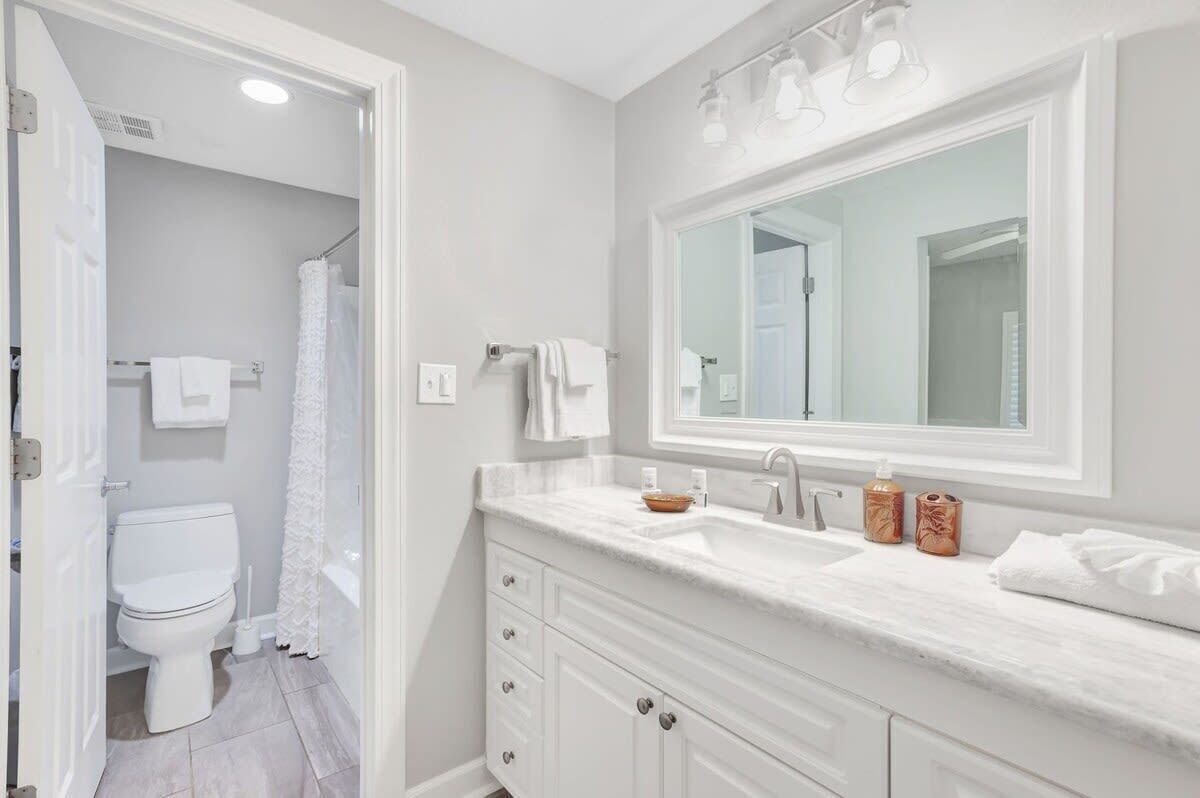 Une salle de bain avec des toilettes blanches, une douche, un grand miroir et un meuble-lavabo avec un évier et deux bouteilles.