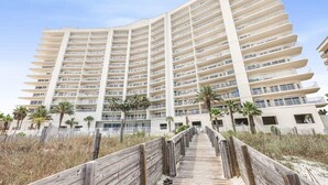 A multi-story hotel with a curved facade, a wooden boardwalk leading to the entrance, and palm trees in the foreground. - Admirals Quarters 802 - Coastal Luxury with Endless Gulf View (Orange Beach)