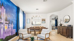 Un soggiorno con un affresco raffigurante un castello, una zona pranzo con un lampadario e una cucina con un armadietto.