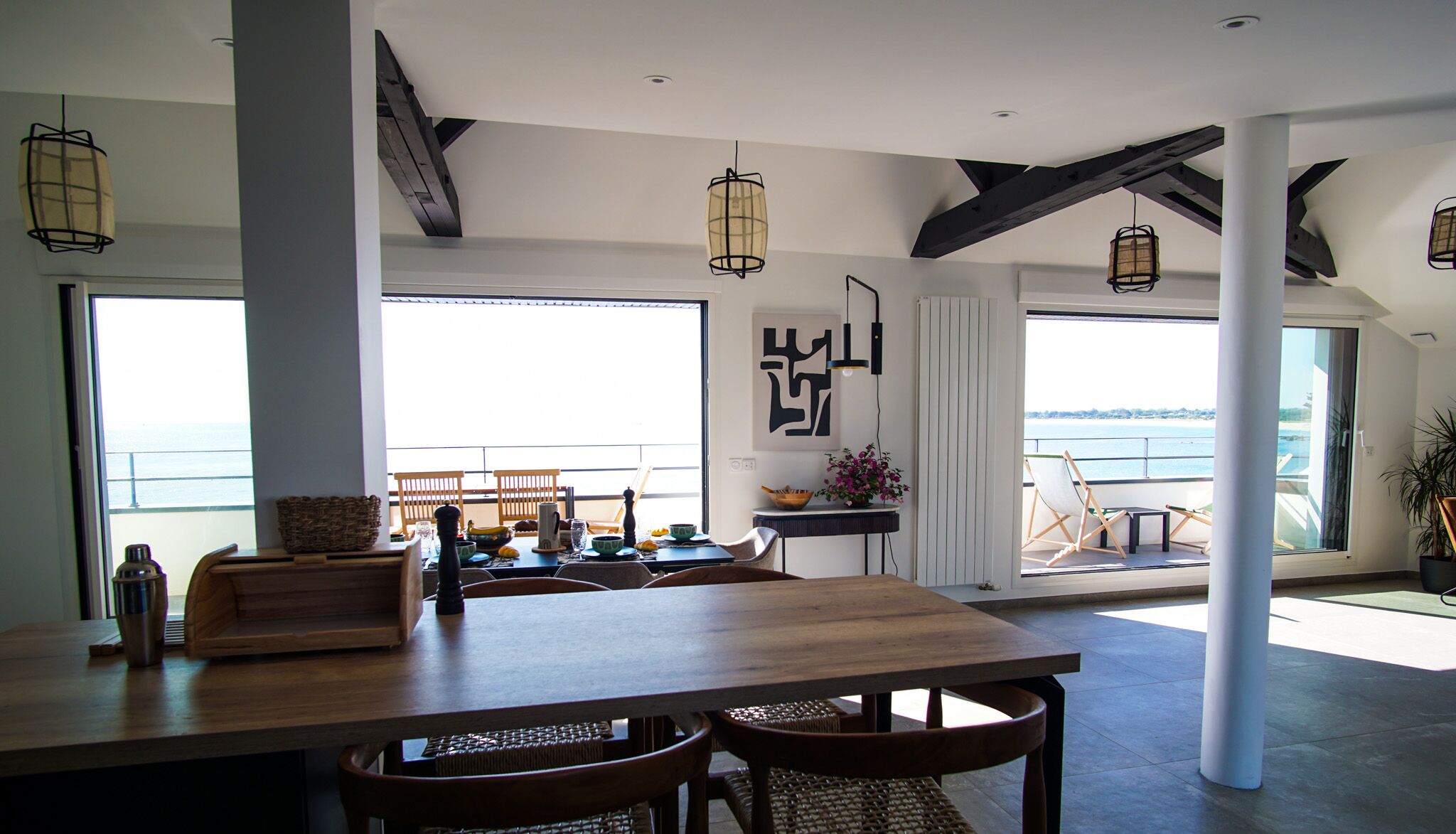 A spacious dining area with a large table and chairs, large windows with a view of the sea, and modern pendant lighting.