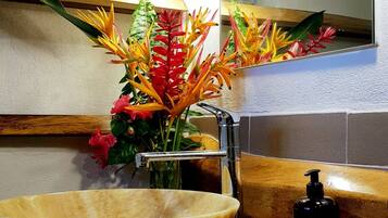 A bathroom with a wooden vanity, a large bowl sink, a mirror with two pendant lights, and a vase of flowers.