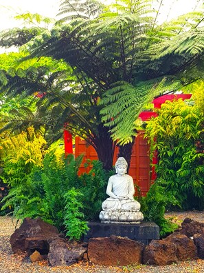 A serene garden with a Buddha statue, lush greenery, and a red building in the background. - Sacred Garden Fiji (Taveuni Island West)