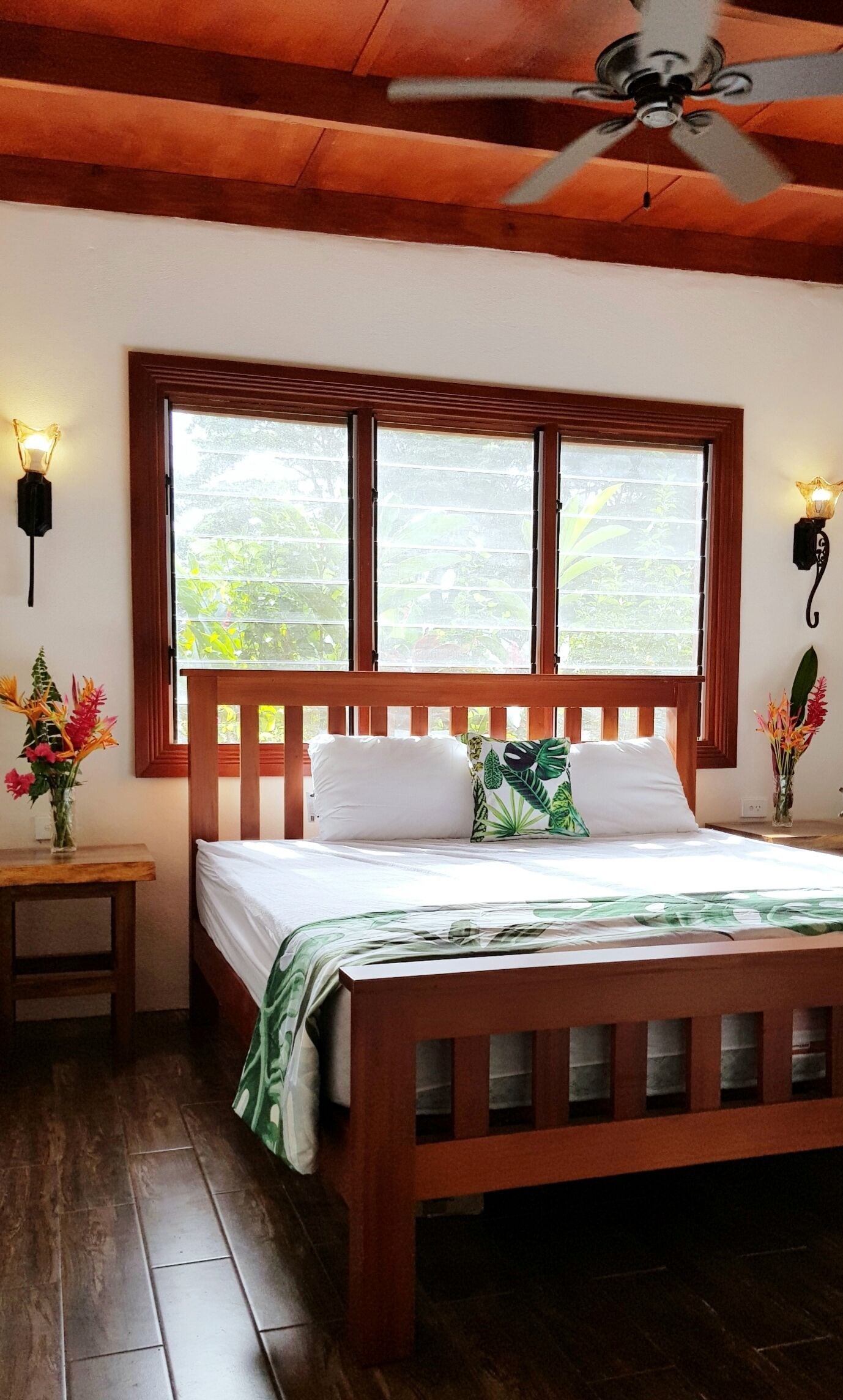 A wooden bed with a view of greenery through a window, a ceiling fan, and a nightstand with a lamp.