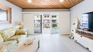A room with a wooden ceiling, a white sofa, a television, a surfboard, and a balcony with a view of buildings.