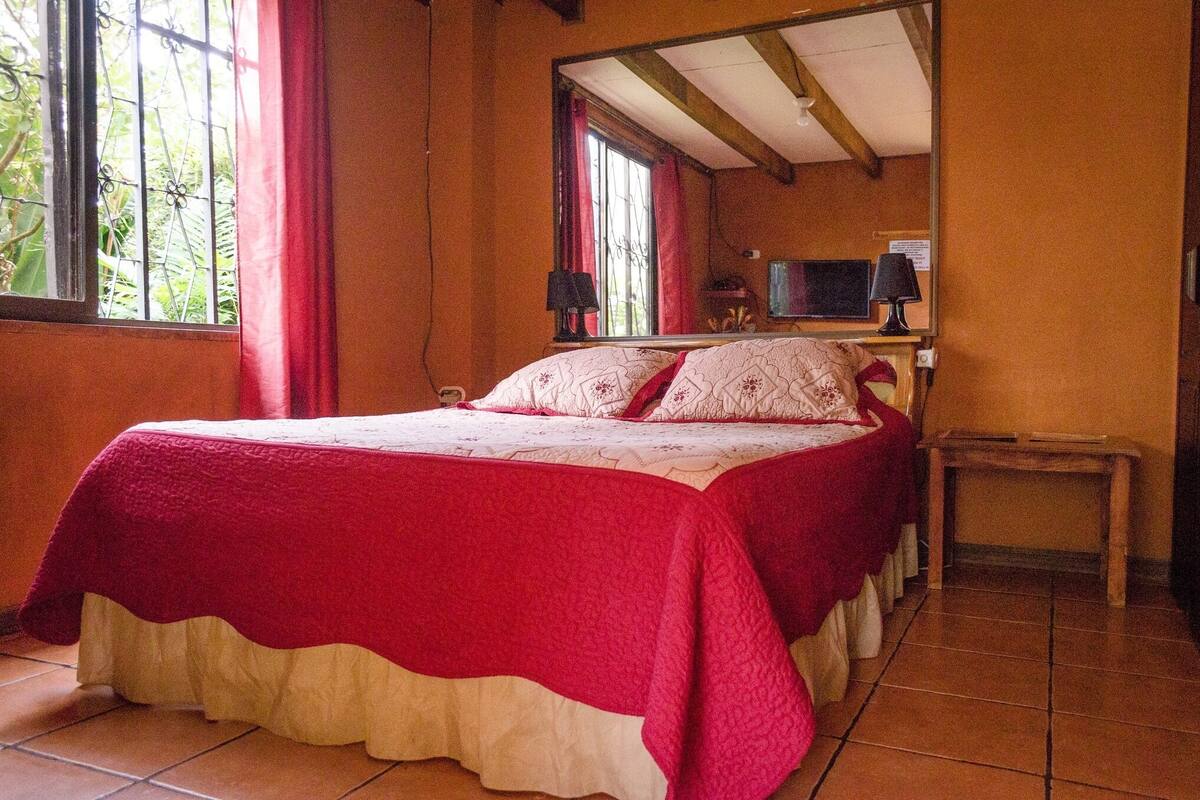 Una cama individual con una colcha roja y blanca, una mesita de noche de madera con una lámpara y una ventana con cortinas rojas.