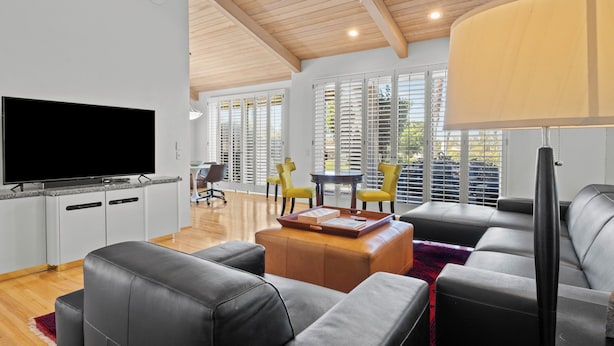 A modern living room with a flat-screen TV, grey leather sofas, a wooden coffee table, and a dining area with yellow chairs.