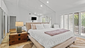 A modern bedroom with a large bed, a wooden nightstand, a TV mounted on the wall, and a window with blinds.