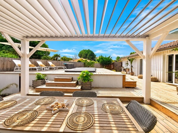 Eine überdachte Terrasse im Freien mit Holzboden, Liegestühlen, einem Esstisch mit gewebten Unterlagen und einer Pergola darüber.
