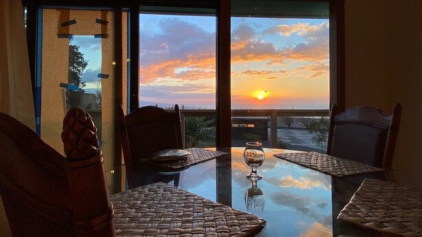 Uma área de refeições com mesa de vidro e jogos americanos trançados, com vista para o pôr do sol através de grandes janelas.