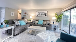 Una sala de estar moderna con un sofá gris, un reposapiés blanco y una mesa de centro. Un cartel en la pared dice "Bienvenido a nuestra Casa de Campo en el Lago".