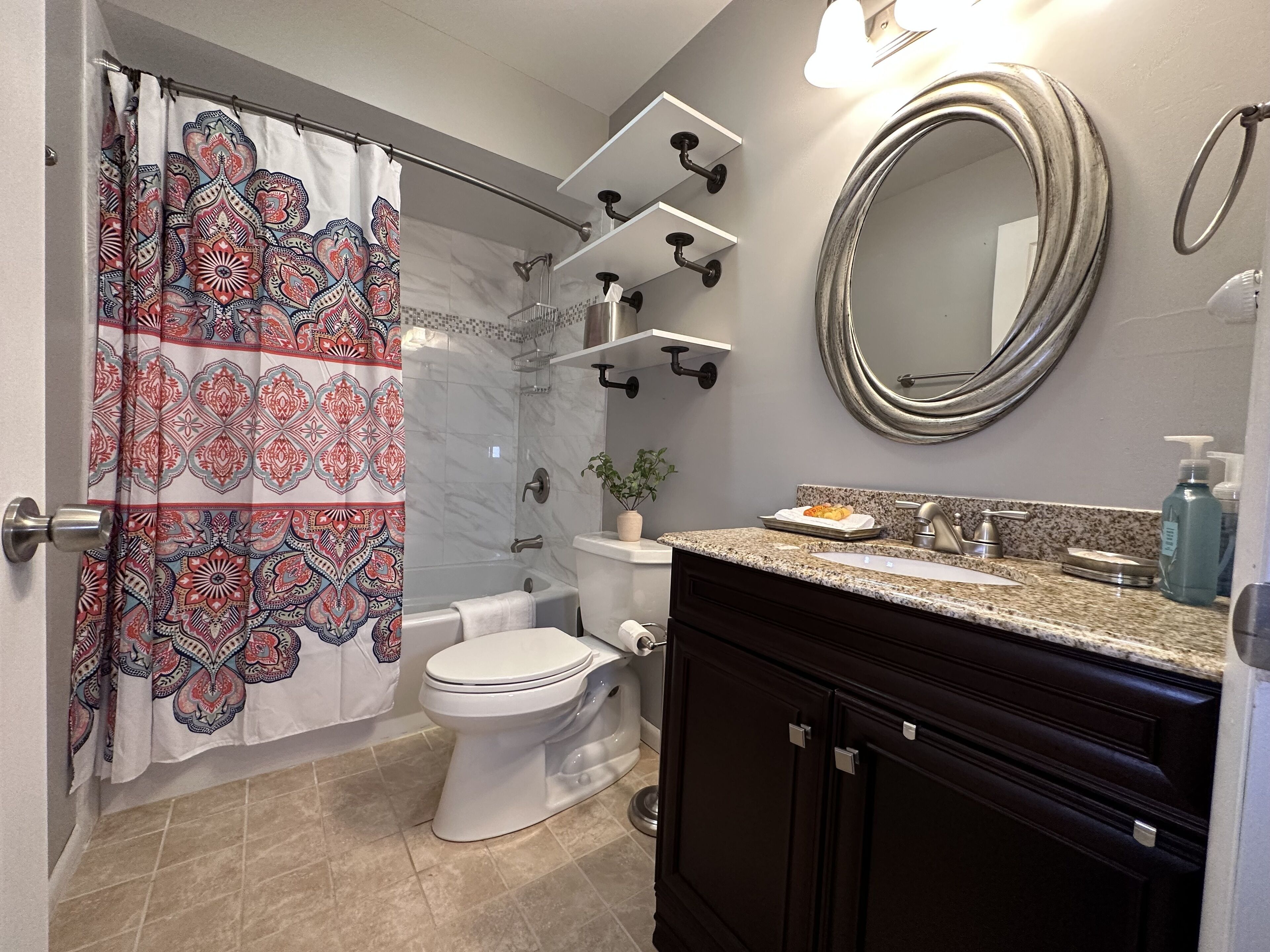 A bathroom with a shower curtain, a toilet, a sink with a mirror, and a shelf with toiletries.