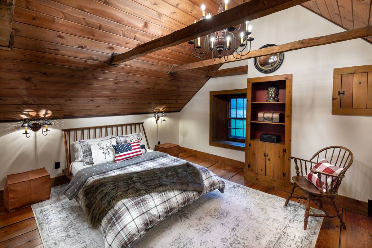 A bedroom with a wooden ceiling, a bed with a plaid bedspread, a rocking chair, and a window with a view.