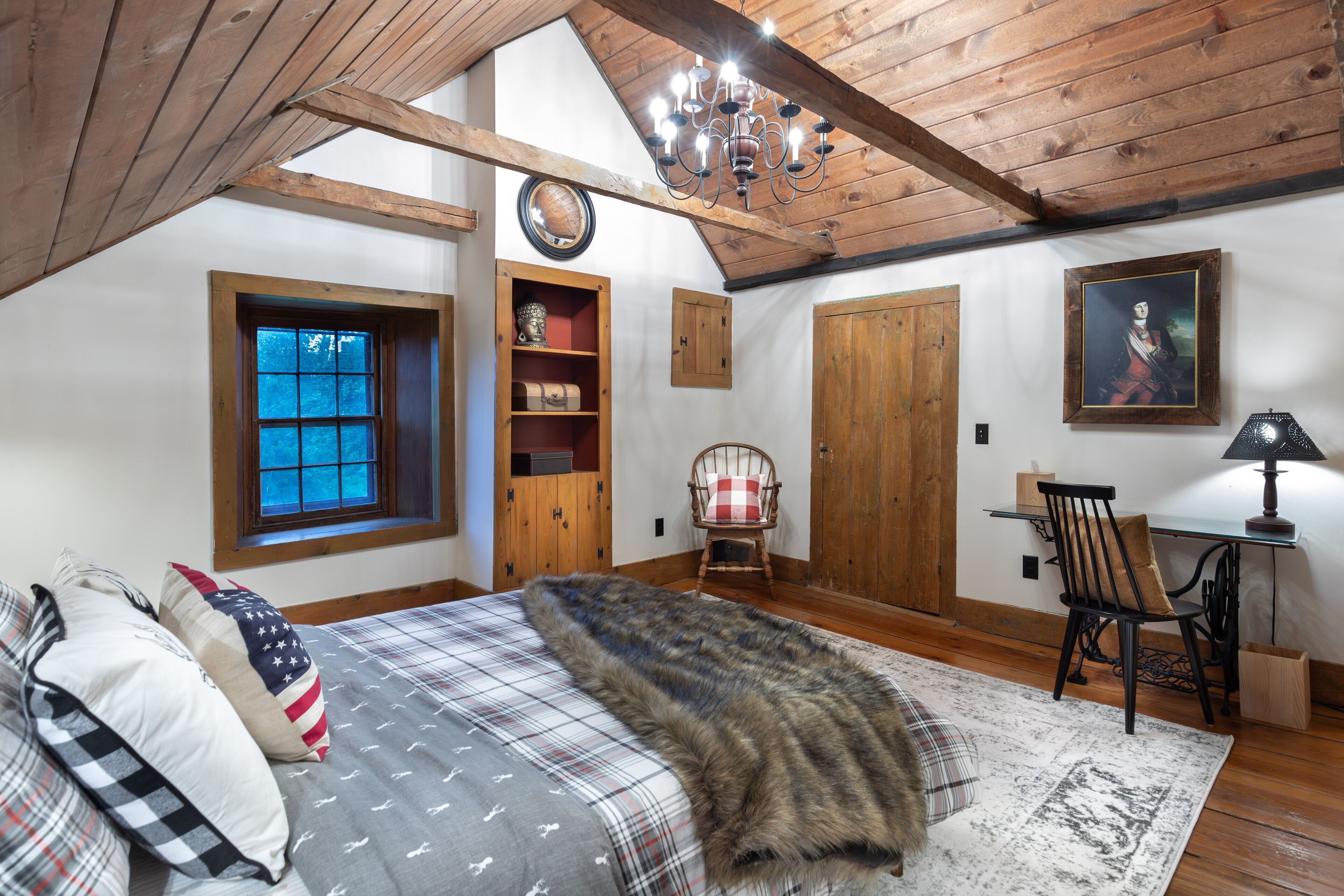 A cozy attic bedroom with a large bed, a wooden ceiling, a window, a chair, and a desk.