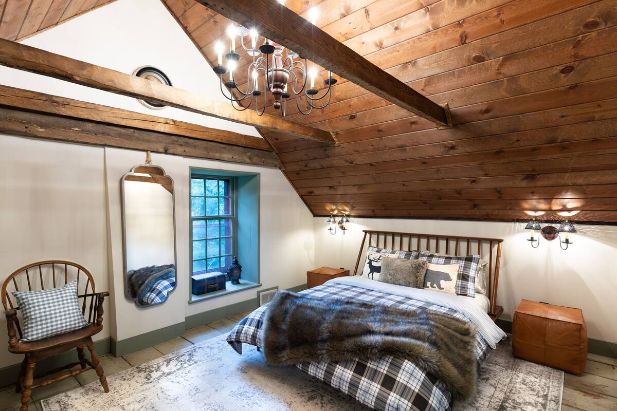 A bedroom with a wooden ceiling, a large bed with a fur throw, a wooden chair, a window with a view, and a chandelier.