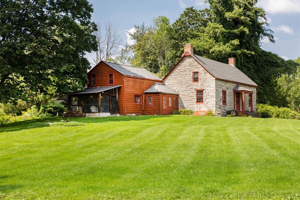 A two-story stone house with a red roof and wooden siding, surrounded by a well-maintained lawn and dense trees.