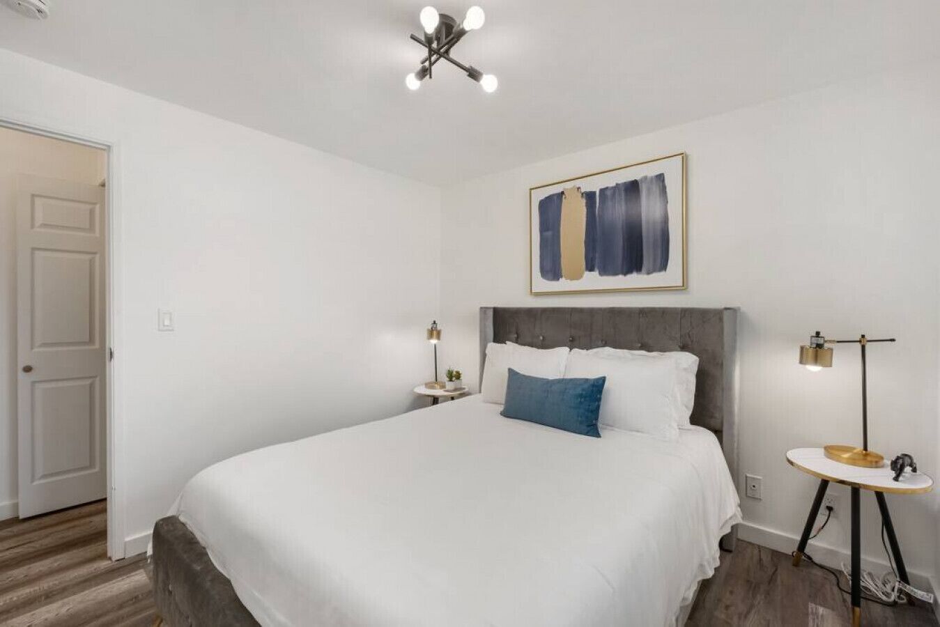 A bedroom with a large bed, a bedside table with a lamp, a small table with a lamp, a door, and a ceiling light fixture.